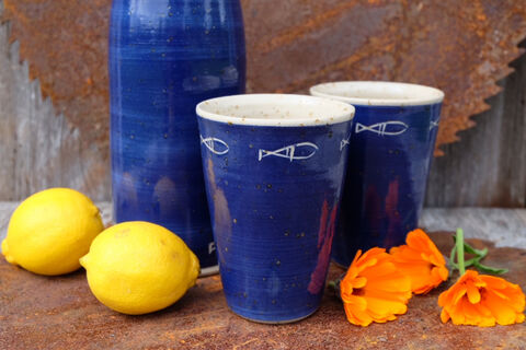 Dunkelblaue Flasche und Becher mit Fischdekor, davor Zitronen und Ringelblumen